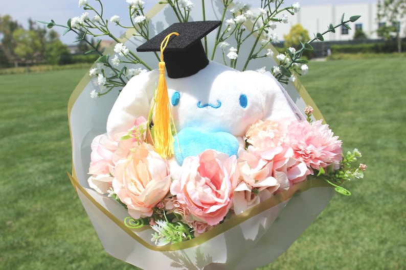 May include: A plush white character wearing a graduation cap, surrounded by pink and peach flowers, and white baby's breath, wrapped in decorative paper. The bouquet has a blue heart-shaped accent and a yellow tassel.