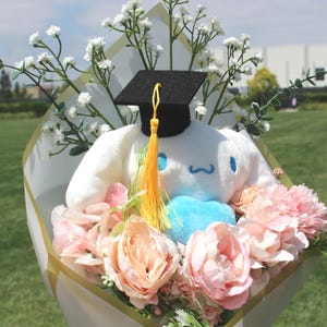 May include: A graduation bouquet featuring a white plush character wearing a black mortarboard with a yellow tassel. The bouquet includes pink and peach-colored flowers, white baby's breath, and a blue heart.