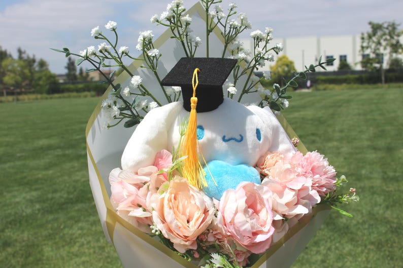 May include: A graduation bouquet featuring a white plush character wearing a black mortarboard with a yellow tassel. The bouquet includes pink and peach-colored flowers, white baby's breath, and a blue heart.