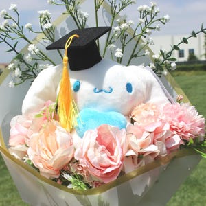 May include: A graduation bouquet featuring a white plush character wearing a black mortarboard with a yellow tassel. The bouquet includes pink and peach-colored flowers, white baby's breath, and greenery, wrapped in decorative paper.