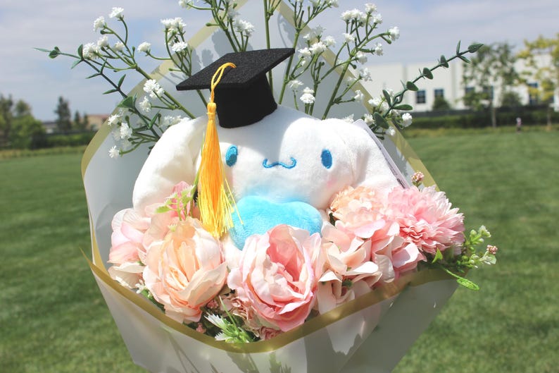 May include: A graduation bouquet featuring a white plush character wearing a black mortarboard with a yellow tassel. The bouquet includes pink and peach-colored flowers, white baby's breath, and greenery, wrapped in decorative paper.