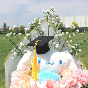 May include: A plush white character wearing a black graduation cap with a yellow tassel, surrounded by pink and peach flowers and white baby's breath. The character has blue accents and is part of a floral bouquet.