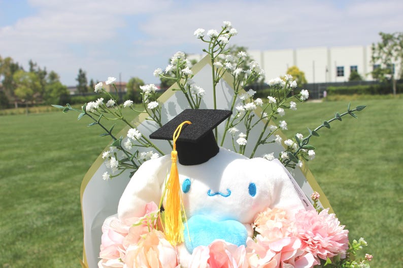 May include: A plush white character wearing a black graduation cap with a yellow tassel, surrounded by pink and peach flowers and white baby's breath. The character has blue accents and is part of a floral bouquet.