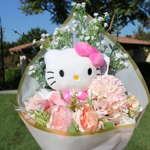 Könnte beinhalten: Ein Strauß aus rosa und weißen Blumen mit einem Hello Kitty Plüschtier in der Mitte. Der Strauß ist in weißes Papier mit einem goldenen Band eingewickelt.