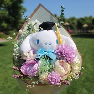 May include: A plush white character with blue accents and a graduation cap, surrounded by a bouquet of pink, purple, and cream-colored flowers. The bouquet is wrapped in a decorative paper.