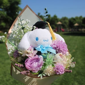 May include: A plush white character with blue eyes and a blue bow tie, wearing a graduation cap, is nestled in a bouquet of purple, pink, and white flowers. The bouquet is wrapped in decorative paper, set against a green background.