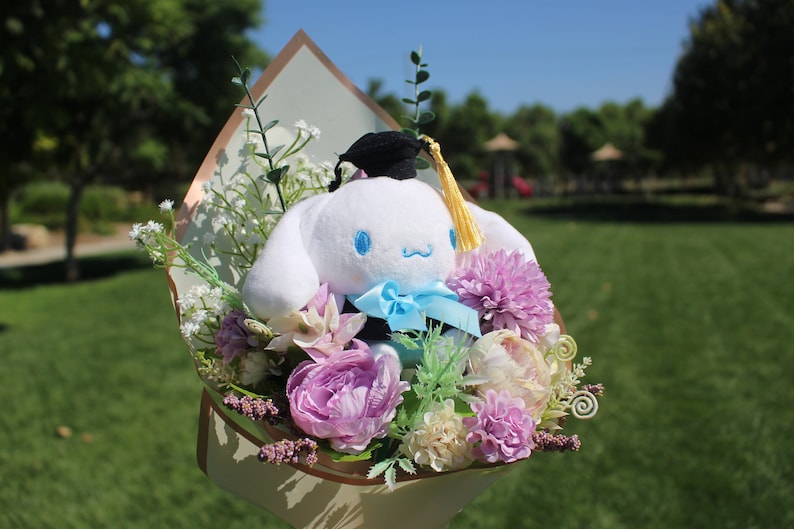 May include: A plush white character with blue eyes and a blue bow tie, wearing a graduation cap, is nestled in a bouquet of purple, pink, and white flowers. The bouquet is wrapped in decorative paper, set against a green background.