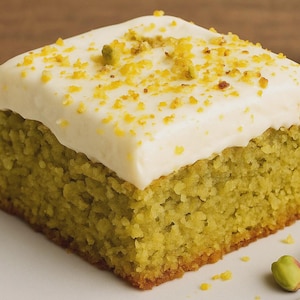 Può includere: Una fetta quadrata di torta al pistacchio con glassa bianca e cosparsa di pistacchi tritati e scorza. La torta è di colore verde chiaro e si trova su un piatto bianco. La torta è un dessert.