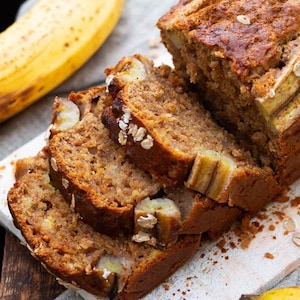 May include: A close-up of a loaf of banana bread sliced into four pieces. The bread is a light brown colour and has a slightly crumbly texture. The slices are stacked on top of each other, and the top slice is slightly tilted. The bread is garnished with oats and banana slices.