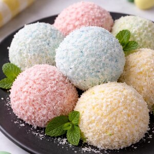 Può includere: Un piatto nero contiene sette dessert rotondi e colorati. Ognuno è ricoperto di cocco grattugiato, con colori tra cui rosa, blu, verde e giallo. Foglie di menta fresca sono sparse intorno ai dessert, aggiungendo un tocco di freschezza.