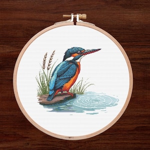 May include: A cross-stitch pattern of a blue and orange kingfisher perched on a branch over a body of water. The bird has a long, pointed beak and is surrounded by green reeds.