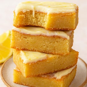 May include: A stack of four lemon bars with a white glaze. The bars are square-shaped with golden-brown edges and a light yellow interior. One bar has a bite taken out of it. A slice of lemon is visible on the side.