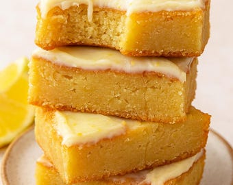 Può includere: Una pila di quattro barrette al limone con glassa bianca. Le barrette sono quadrate con bordi dorati e un interno giallo chiaro. A una barretta manca un morso. Una fetta di limone è visibile.