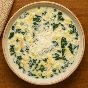 以下が含まれることがあります： ベージュのボウルに入った、ほうれん草とパスタのクリーミーなスープ。スープにはすりおろしたチーズと黒胡椒がトッピングされています。ボウルは木製の表面に置かれ、横にはベージュの布があります。