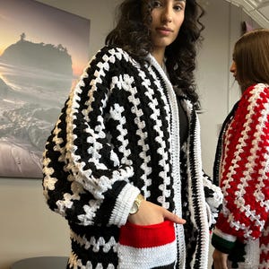 May include: A black and white crocheted cardigan with a red, white and black pocket detail. The cardigan has a loose fit and long sleeves.