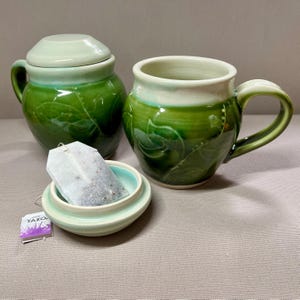May include: A green ceramic teacup and lidded jar with a leaf design. The teacup has a white rim and a handle. The jar has a white lid. A tea bag is in the lidded jar.