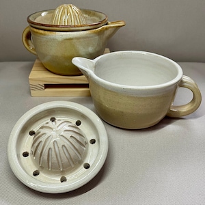 May include: A ceramic citrus juicer set with a light beige and brown glaze. The set includes a juicer, a pitcher with a spout and handle, and a separate reamer. The juicer sits on a small wooden stand.