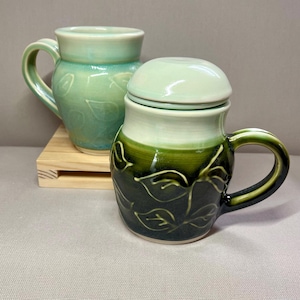 May include: Two ceramic mugs in shades of green. One is a light green mug with leaf imprints. The other is a darker green mug with a lid, also with leaf imprints. Both mugs have handles and are on a small wooden stand.
