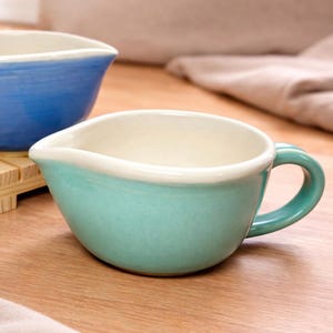 May include: A turquoise ceramic bowl with a handle and a pouring spout. The interior is off-white, and the rim is trimmed with white. Another blue bowl is visible in the background. The bowls are on a wooden surface.