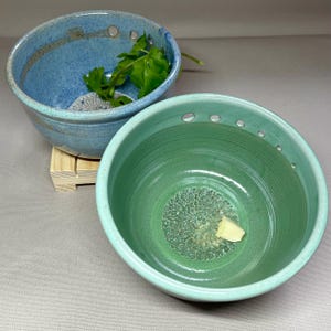 May include: Two ceramic bowls with drainage holes. The bowl on the left is blue and white with green herbs inside. The bowl on the right is green with a white piece of soap inside.