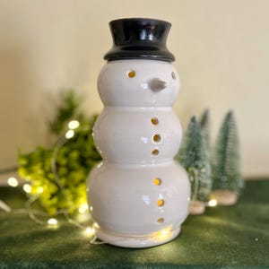 May include: A ceramic snowman-shaped oil warmer with a glossy white finish and a black top hat. The snowman has a carrot nose and circular cutouts for light to shine through. Decorative holiday item.