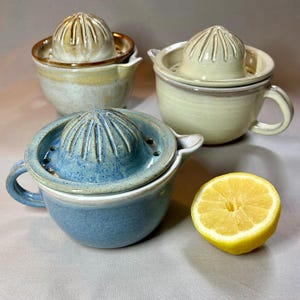 May include: Three ceramic citrus juicers in blue, beige, and cream. Each has a ridged reamer and a small handle. A halved lemon is next to the blue juicer. The handmade juicers have a rustic, artisanal appearance.