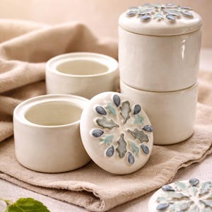 May include: Cream-colored ceramic jars with lids, featuring a raised floral design in shades of blue and gray. The jars are cylindrical and stacked, with a lid off to the side, revealing the interior. The set is displayed on a neutral-toned cloth.