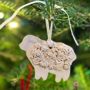 May include: A white ceramic sheep ornament with a textured, bumpy surface on its body representing wool. The ornament has a smooth head and legs, a hole at the top for hanging, and is suspended by a thin white ribbon tied in a simple loop.
