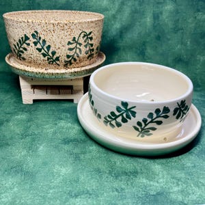 May include: Two ceramic planters with matching saucers. The larger planter is speckled beige with green leaf designs. The smaller planter is white with green leaf designs. Both planters are suitable for indoor or outdoor use.