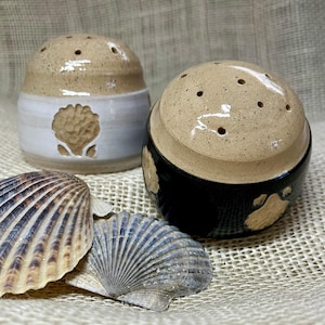 May include: Two ceramic salt and pepper shakers with a speckled tan top and a white and black base. The white shaker has a seashell design. Seashells are in the foreground.
