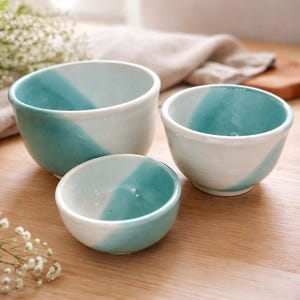 May include: Three ceramic bowls in varying sizes, each with a diagonal split of white and teal glaze. The bowls are arranged on a wooden surface, with a soft, blurred background. The bowls are likely for serving or decorative use.