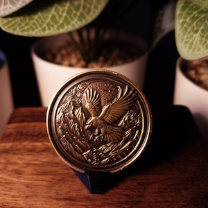 May include: A gold-toned metal coin with a detailed design of a bird in flight. The coin is on a wooden surface.