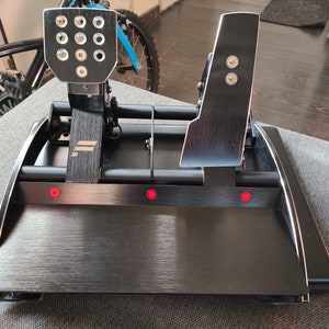 Peut inclure: Un ensemble de pédales de course noir et argenté avec une finition en métal brossé. L'ensemble comprend une pédale d'accélérateur, une pédale de frein et un repose-pieds. Les pédales ont une surface texturée et sont montées sur une base noire.