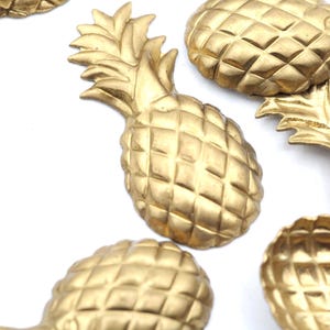May include: Close-up of several gold-colored pineapple-shaped decorative objects. The pineapples have a textured, diamond-patterned surface and detailed leafy tops. The objects are likely made of metal and are arranged against a white background.