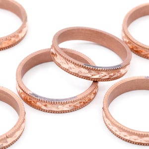May include: Close-up of several copper-colored rings with a textured, geometric pattern around the band. The rings are arranged on a white surface, showcasing their intricate design and metallic sheen. The rings appear to be jewelry.