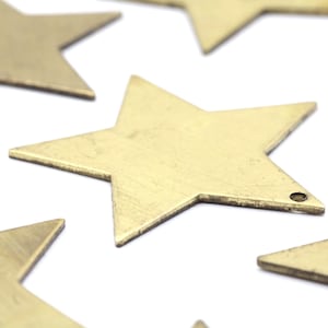 May include: Close-up of several brass star-shaped charms scattered on a white surface. Each star has five points and a small hole for hanging. The stars have a slightly aged, vintage appearance.