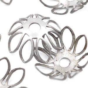 May include: Close-up of several silver-colored metal bead caps. Each cap has a flower-like design with multiple petal-shaped cutouts and a central hole. The caps are scattered against a white background, suggesting a craft or jewelry-making theme.