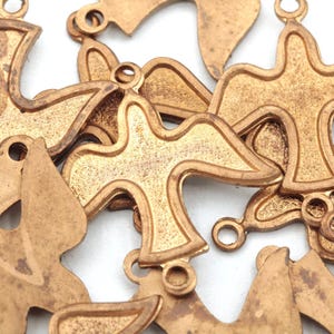 May include: Close-up of several bronze-colored dove charms. Each charm has a detailed outline of a dove with a small loop at the top for hanging. The charms are scattered against a white background, creating a textured effect.