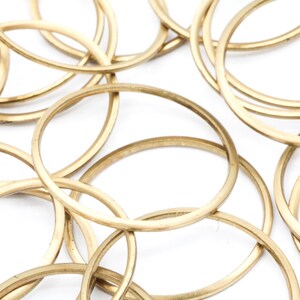 May include: Overhead shot of multiple gold-colored metal rings, overlapping and scattered against a white background. The rings are oval-shaped and appear to be made of a matte finish metal. The image is well-lit, highlighting the texture and shape of the rings.