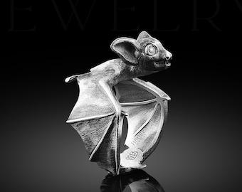 May include: A silver bat ring with detailed wings and a small red eye. The ring is on a black background and reflects in the surface.