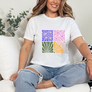 May include: A white t-shirt with a graphic design of four colorful squares featuring stylized flowers. The squares are arranged in a 2x2 grid.