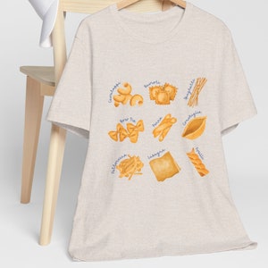 May include: A beige t-shirt with a graphic of different types of pasta, including spaghetti, ravioli, lasagna, and bow tie pasta. The names of each pasta type are written in blue text.