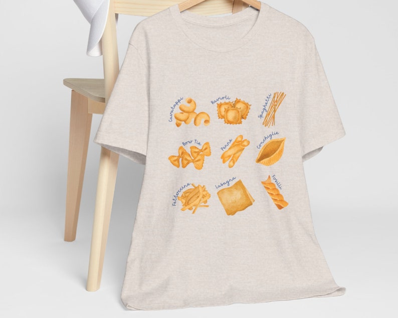 May include: A beige t-shirt with a graphic of different types of pasta, including spaghetti, ravioli, lasagna, and bow tie pasta. The names of each pasta type are written in blue text.