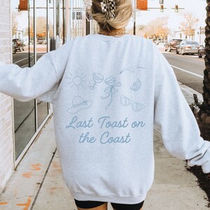 May include: A light gray sweatshirt with a blue graphic design of a sun, two glasses of wine, a bow, a lemon slice, sunglasses, and a hat. The text "Last Toast on the Coast" is printed below the graphic.