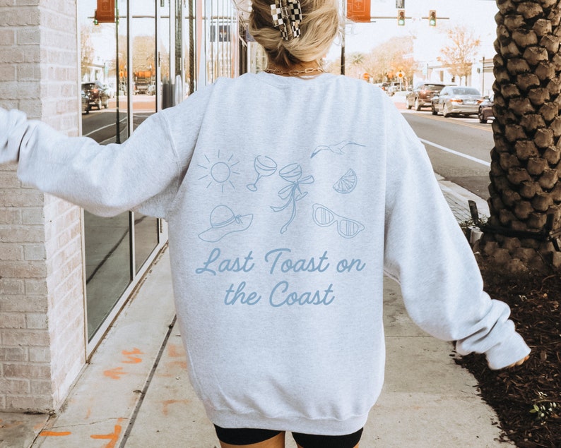 May include: A light gray sweatshirt with a blue graphic design of a sun, two glasses of wine, a bow, a lemon slice, sunglasses, and a hat. The text "Last Toast on the Coast" is printed below the graphic.