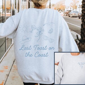May include: Gray sweatshirt with a blue graphic of a sun, sunglasses, a hat, a glass of wine, a lemon, and a seagull. The text "Last Toast on the Coast" is printed in blue below the graphic. The sweatshirt is shown on a model and a close-up of the front of the sweatshirt is shown.