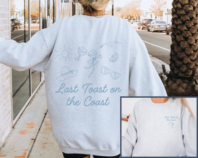 May include: Gray sweatshirt with a blue graphic of a sun, sunglasses, a hat, a glass of wine, a lemon, and a seagull. The text "Last Toast on the Coast" is printed in blue below the graphic. The sweatshirt is shown on a model and a close-up of the front of the sweatshirt is shown.