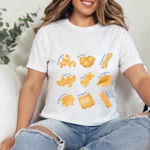 May include: A white t-shirt with a graphic of different types of pasta, including spaghetti, lasagna, tortellini, and more. Each pasta type is labeled with its name in blue text.