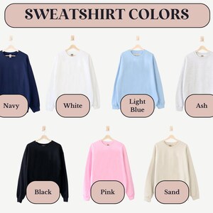 May include: A display of eight sweatshirts in different colors, including navy, white, light blue, ash, black, pink, and sand. The sweatshirts are hanging on hangers against a white background. The text "SWEATSHIRT COLORS" is displayed above the sweatshirts.