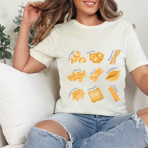 May include: A white t-shirt with a graphic of different types of pasta, including spaghetti, tortellini, and lasagna. Each pasta type is labeled with its Italian name.